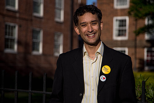 Dave Raval smiling and looking straight ahead. Blocks of redbrick flats in the background